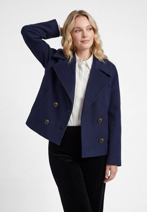 Blonde woman wearing a navy pea coat over a white shirt and black pants, with one hand behind her head, looking to the side.