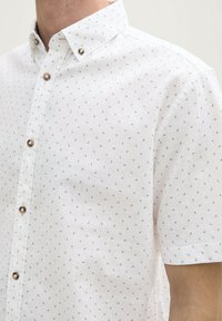 Chemise à manches courtes en tissu blanc avec un motif à pois bleus. Dotée d'un col pointu et de boutons marron brillants.