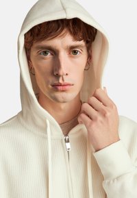 Sudadera blanca con canalé, con cremallera frontal y capucha con cordón. El modelo ajusta la capucha, mostrando una textura suave y un diseño limpio.