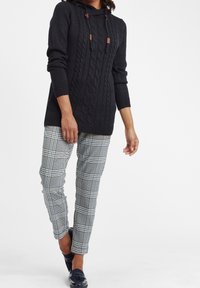 Zwarte gebreide trui met kabelpatroon en capuchon, gecombineerd met een geruite broek in blauw, zwart en wit. Platte schoenen maken de outfit compleet.