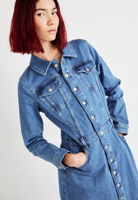 Denimkjole med knappelukking foran, krage, to brystlommer og lange ermer. Materialet er en middels blå vask med en lett falmet tekstur.