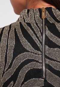 Czarny top z tkaniny z błyszczącym złotym wzorem w paski zebry, wysoki kołnierz oraz widoczny metalowy zamek z tyłu.