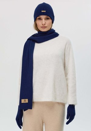 Set di cappello e sciarpa a coste color navy, abbinato a un maglione bianco a trama. I pantaloni di un delicato beige completano il look. La sciarpa si drappeggia elegantemente, i guanti sono coordinati.