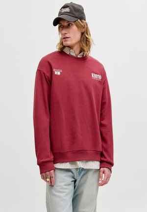 Jeune personne aux cheveux mi-longs porte une casquette noire, un sweat rouge avec le texte « Kyoto », une chemise rayée en dessous et un jean bleu clair.