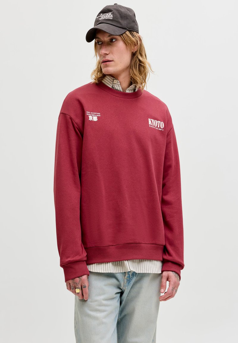 Jeune personne aux cheveux mi-longs porte une casquette noire, un sweat rouge avec le texte « Kyoto », une chemise rayée en dessous et un jean bleu clair.