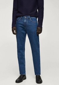 Jeans de mezclilla azul oscuro con corte recto, que cuentan con cinco bolsillos y una cremallera. Acompañados de un suéter de punto azul marino y zapatos negros.