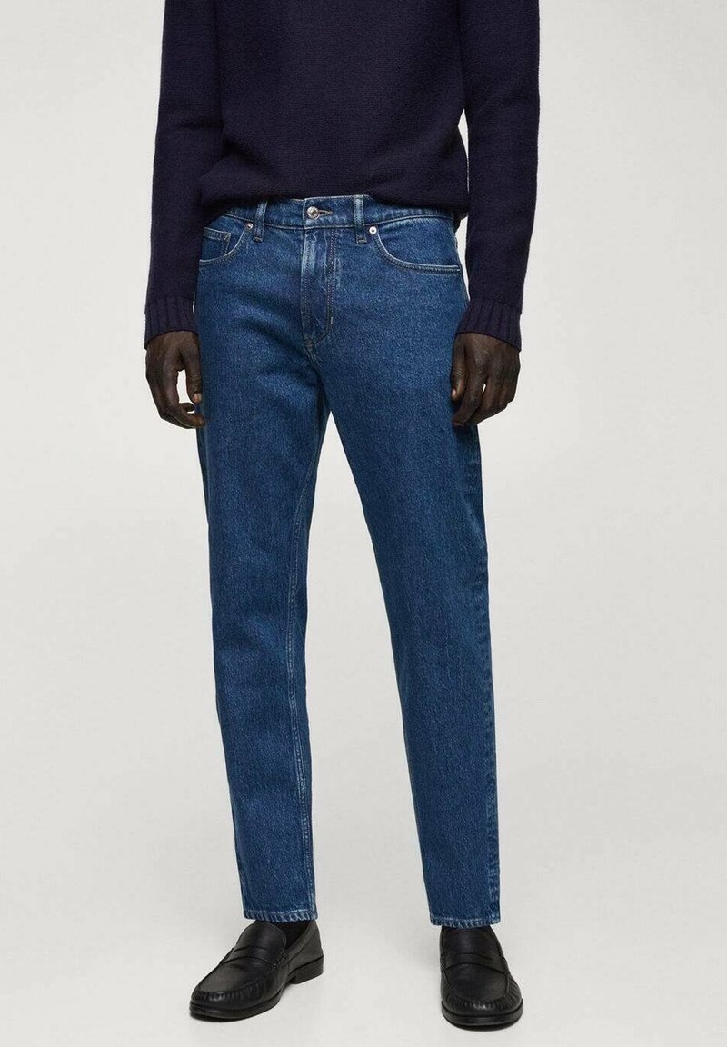 Jeans de mezclilla azul oscuro con corte recto, que cuentan con cinco bolsillos y una cremallera. Acompañados de un suéter de punto azul marino y zapatos negros.
