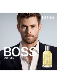 Le parfum BOSS Bottled dans un flacon en verre transparent avec un liquide jaune. Un mannequin portant un costume sombre et une chemise blanche, avec une barbe bien entretenue.