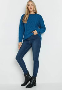 Blauer Kabelstrickpullover mit langen Ärmeln und Schulterknöpfen, kombiniert mit dunklen Skinny-Jeans und schwarzen Knöchelstiefeln mit Hardware-Details.
