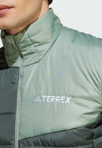 Giacca puffer verde con colletto alto, dotata di chiusura con zip e logo ricamato "TERREX" in bianco. Texture liscia con finitura opaca.