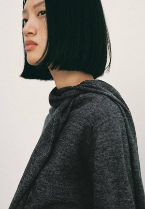Joven con cabello negro liso en un corte bob, vistiendo un suéter con capucha gris oscuro, mirando hacia la izquierda sobre un fondo liso.
