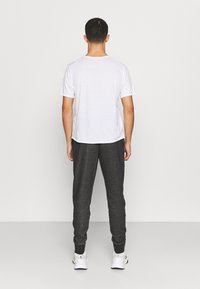 Weißes Kurzarm-T-Shirt kombiniert mit dunkelgrauen Jogginghosen mit elastischen Bündchen und weißen Sportschuhen. Rückansicht vor einem einfarbigen Hintergrund.