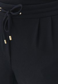 Pantaloni de trening bleumarin cu talie reglabilă, prezentând accente din metal aurit și un material moale, texturat. Design plisat în față.