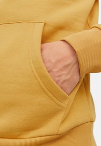 Gelbe Sweatshirt-Stoff mit weicher Textur, ausgestattet mit einer Fronttasche und gerippten Bündchen. Sichtbare Hand, die in der Tasche ruht.