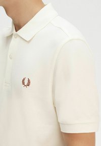 Polo bianco realizzato in tessuto strutturato, con patta a due bottoni e logo ricamato marrone sul petto. Maniche corte.
