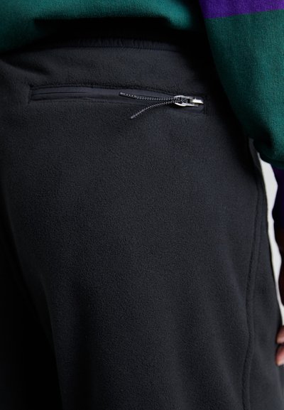 Calças de polar pretas com um bolso com fecho nas costas. O tecido parece macio, com uma textura suave e um ajuste justo na cintura.