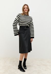 Schwarz-weiß gemusterter Pullover mit weiten Ärmeln, kombiniert mit einem einfarbigen schwarzen knielangen Rock mit seitlicher Tasche und Gürtel. Schwarze Schuhe mit Absatz.