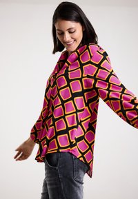 Langarmbluse aus glänzendem Stoff, mit einem pinken und gelben geometrischen Muster auf schwarzem Hintergrund. Lockerer Schnitt mit abgerundetem Saum.