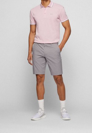 Mann trägt hellrosa kurzärmliges Poloshirt, graue knielange Shorts, weiße Sportsocken und weiße Turnschuhe, steht mit einer Hand in der Hosentasche.