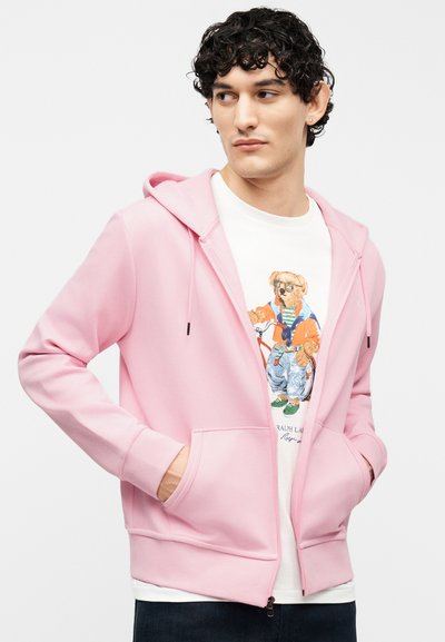 Νεαρός με σγουρά σκούρα μαλλιά που φοράει ένα ανοιχτό ροζ φούτερ με φερμουάρ πάνω από ένα λευκό T-shirt με μοντέρνο σχέδιο αρκούδας.