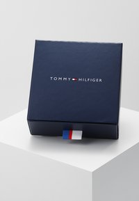 Ambalaj pătrată de culoare albastru marin cu un finisaj lucios, având "TOMMY HILFIGER" scris cu litere albe și un accent dungi albastre, albe și roșii pe latură.