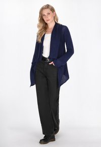 Donna che indossa un cardigan aperto blu navy, una maglietta bianca, jeans larghi neri e stivali neri con lacci su uno sfondo bianco.