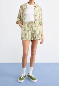 Zelená košile s krátkým rukávem a sladěné šortky s růžovými květinovými vzory, dřevěnými knoflíky a elastickým pasem. Na nohou zelené tenisky.