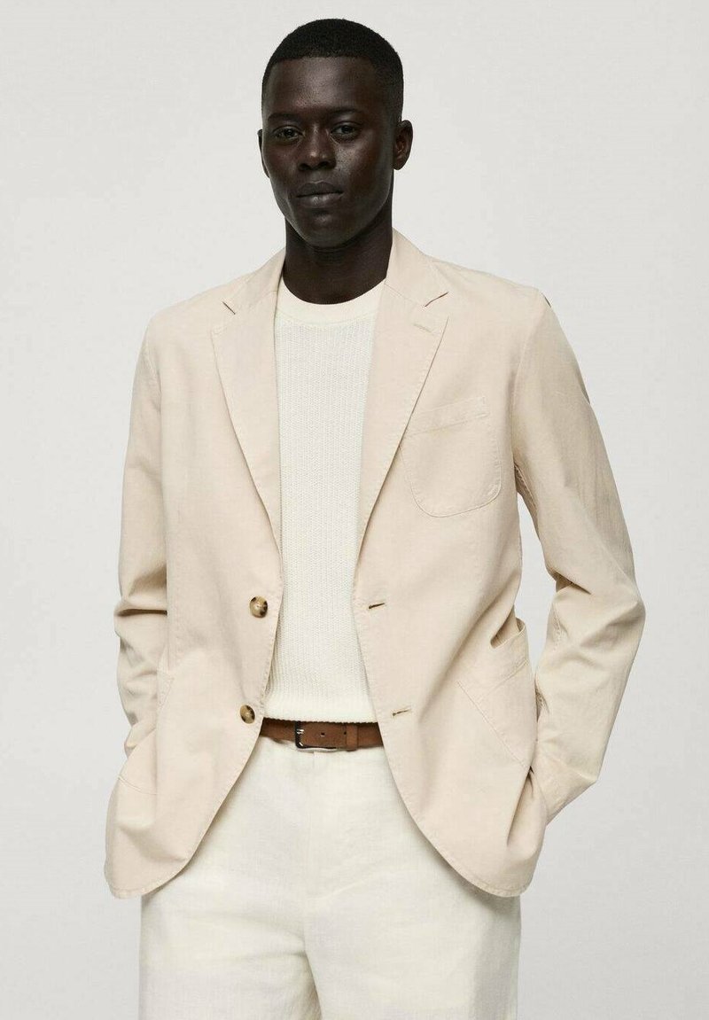 Blazer beige in lino con due bottoni, rever a punta e taschino sul petto, indossato sopra un maglione bianco in maglia e pantaloni chiari.