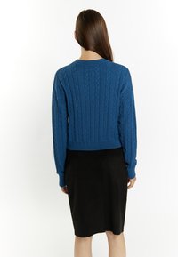 Maglione blu a treccia con un design corto e polsini a costine, abbinato a una gonna nera aderente. La vista da dietro evidenzia la texture e il contrasto di colori.