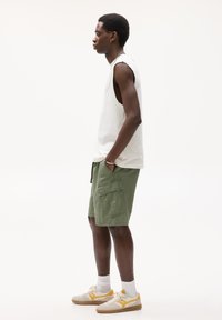 T-shirt blanche sans manches, shorts cargo verts avec poches à rabat, et baskets beiges avec accents jaunes. Design simple et décontracté.