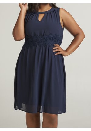 Robe bleu marine sans manches avec encolure en clé et détail de ceinture en dentelle, portée par une personne avec une main sur la hanche et l'autre levée.