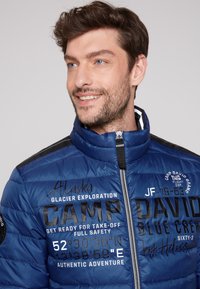 Giacca puffer blu con colletto alto, chiusura con zip e dettagli/lettere nere. Il testo include coordinate geografiche e loghi del marchio.
