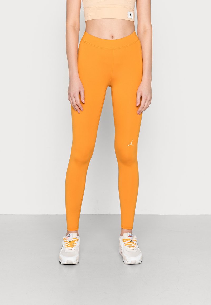 Orange leggings med hög midja och en slät yta, med en liten vit logotyp. Kombinationen med vita sneakers med orange detaljer.