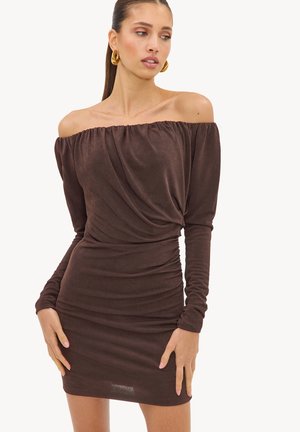 Femme portant une robe marron cintrée à manches longues et épaules dénudées avec des détails froncés, posant les mains près des hanches et regardant sur le côté.