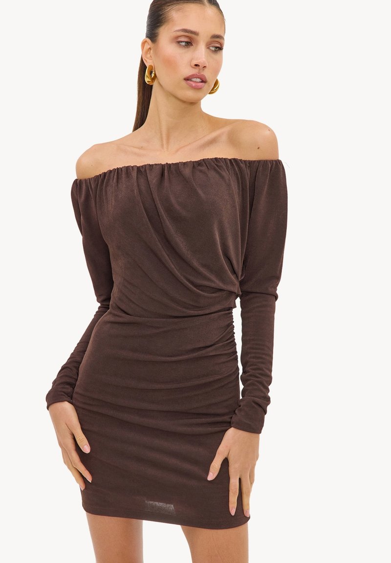 Femme portant une robe marron cintrée à manches longues et épaules dénudées avec des détails froncés, posant les mains près des hanches et regardant sur le côté.