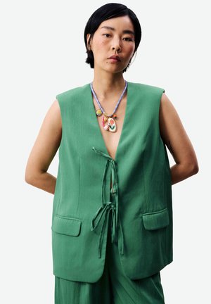 Femme aux cheveux courts noirs portant un gilet vert sans manches noué devant et un collier pendentif coloré, debout les mains derrière le dos.