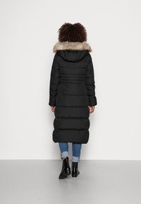 Abrigo acolchado negro con capucha forrada de piel, diseño acolchado, largo y con bolsillos laterales, combinado con jeans azules remangados y botas negras de tobillo.