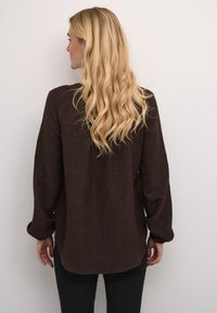 Chemise marron à manches longues avec un tissu texturé, coupe décontractée et poignets froncés, associée à un pantalon noir. Vue arrière sur fond uni.