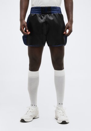 Άνδρας που φοράει μαύρα αθλητικά σορτς με μπλε μέση, λευκές κάλτσες μέχρι το γόνατο και λευκά αθλητικά παπούτσια, στέκεται μπροστά σε απλό φόντο.