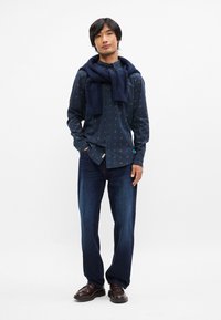 Μπλε σχέδιο πουκάμισο με κουμπιά, σκούρα τζιν, μπλε πλεκτός σκαφάκι ριγμένος στους ώμους, καφέ loafers, στέκεται μπροστά σε λευκό φόντο.