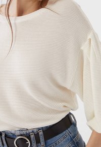 Suéter de canalé en color crema con un amplio cuello redondo y mangas cortas, combinado con un cinturón negro, usado sobre unos pantalones vaqueros. Textura suave.