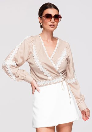 Vrouw met grote bruine zonnebril, beige wikkelblouse met witte kanten details en witte minirok tegen een effen achtergrond.