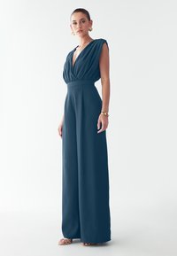 Teal värvi ilma varrukateta kombinesoon sügava V-kaelusega ja pleediga bodi; sellel on sobiv vöökoht ja laiad püksid; sile, mati tekstuur.