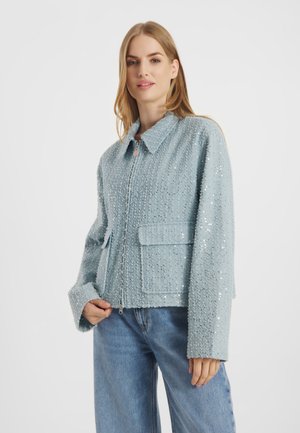 Kobieta ubrana w jasnoniebieską, teksturowaną kurtkę na zamek z klapkowymi kieszeniami i szerokimi rękawami, zestawioną z niebieskimi dżinsami, stojąca na jednolitym tle.