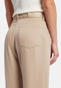 Beige jeans met een gestructureerde stof, zichtbare achterzakken en een leren merklabel op de tailleband. Tailored fit en schone lijnen.