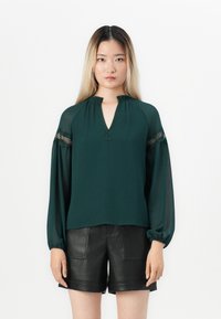 Smaragdgrønn bluse med V-hals, samlet detalj og gjennomsiktige ermer. Har teksturerte detaljer langs halsen og ermene.