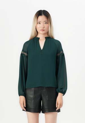 Emerald green blouse with a V-neckline, gathered detail, and sheer sleeves. Features textured accents along the neckline and sleeves.