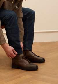 Bruine leren enkelboots met vetersluiting en gouden oogjes, gedragen met blauwe spijkerbroek op een houten vloer.