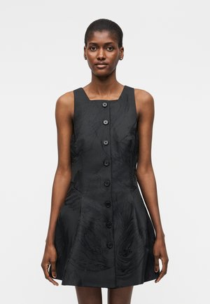 Γυναίκα με μαύρο αμάνικο φόρεμα με κουμπιά μπροστά και διακριτικό υφασμάτινο λουλουδάτο μοτίβο, στέκεται μπροστά σε απλό ανοιχτό γκρι φόντο.