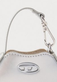 Detailní záběr bílé rukojeti kabelky s viditelným prošitím, stříbrným zapínáním a oválným raženým logem na hladkém materiálu.